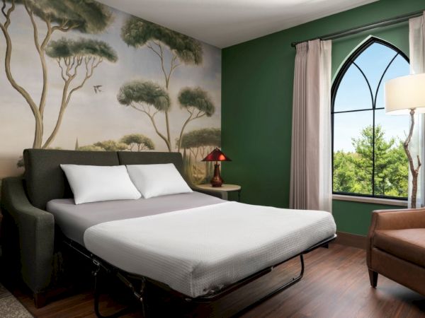 A hotel bedroom with a mural of trees on the wall, a grey bed with white linens, green accent wall, large arched window, and a brown armchair nearby. Top it at 140 characters, always ending the sentence.