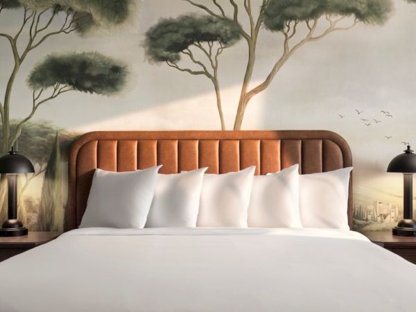 A cozy hotel bed with a padded headboard, white linens and multiple pillows, flanked by matching nightstands and lamps, fronted by a nature mural.