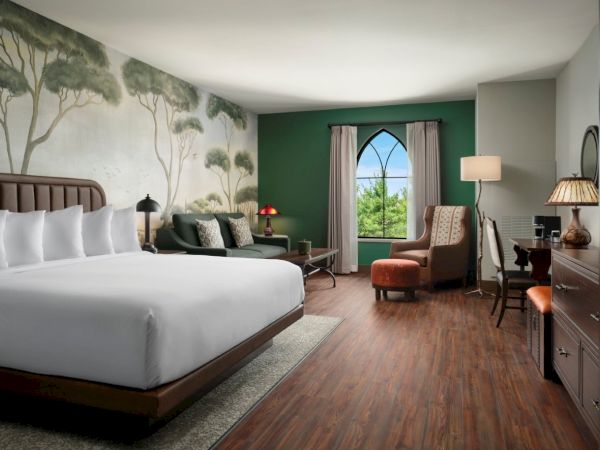 A hotel room with a large bed, nature mural, green accent wall, armchairs, a desk, and tree-like wallpaper, offering a calm, spacious ambiance.