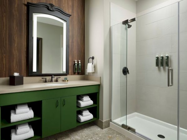 A modern bathroom with a green vanity, open shelves with towels, a dark-framed mirror, and a glass-walled shower.