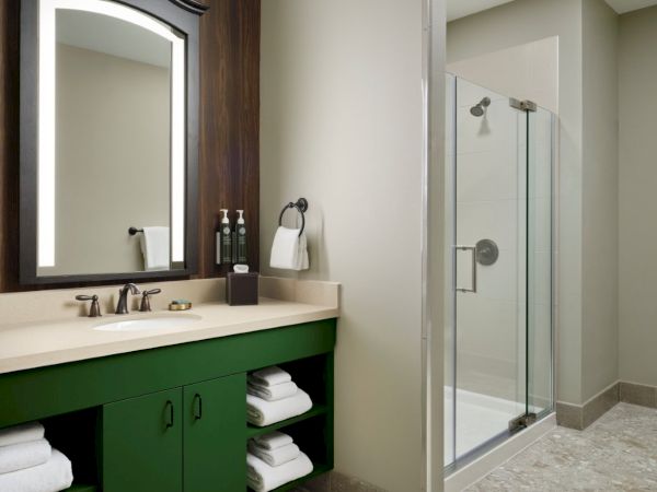 A modern bathroom with a green vanity, white countertop, two towels on shelves, a large framed mirror, and a glass-walled shower.