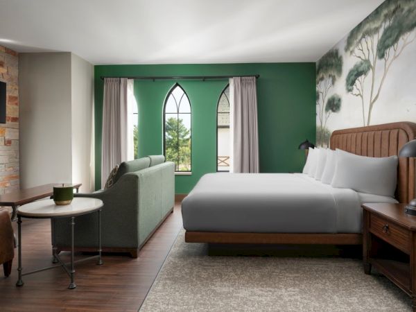 A modern hotel room with a green accent wall, a large bed, a green sofa facing glass doors, and a tree mural, creating a calm, nature-inspired vibe.