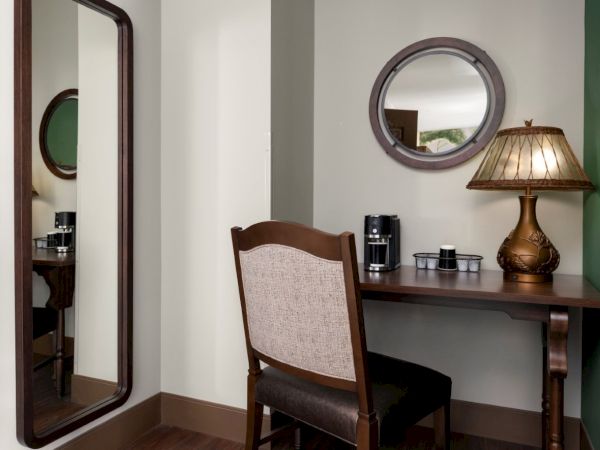 A small hotel-style desk setup with a wooden chair, a round mirror, a table lamp, and a coffee/tea station against a wall.