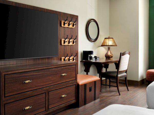 A hotel room with a large flat-screen TV, a dresser, a built-in wine rack, a small desk and chair, a side lamp, and a bed nearby.
