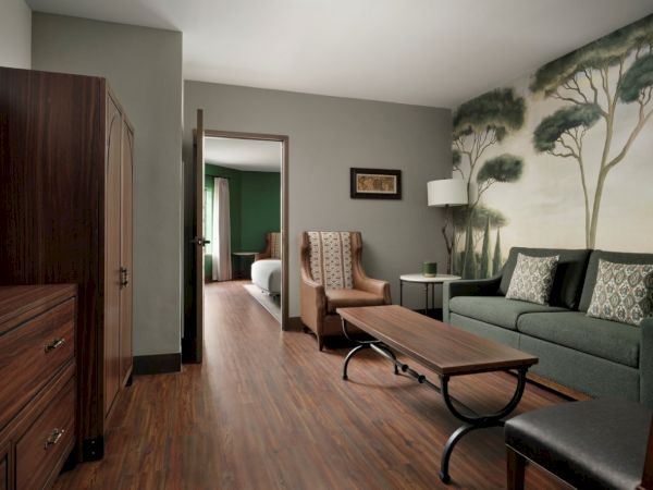A cozy living room with a green sofa, wooden coffee table, tall cabinet, and a mural of trees on the wall, leading to a bedroom in the back.