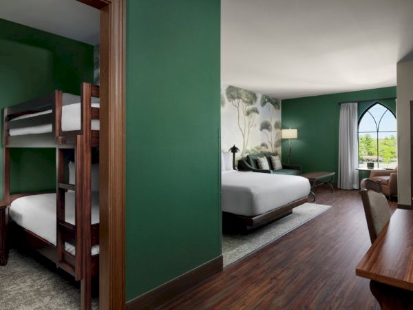 A hotel room with a green accent wall, a doorway to bunk beds on the left, a large bed and seating area on the right, and a window with a view in the distance.