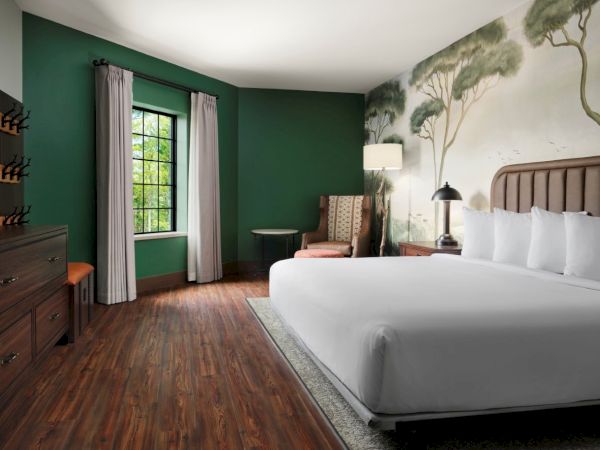 A stylish hotel room with a large bed, dark green walls, wooden floor, a mural of trees above the headboard, and a seating area by the window.