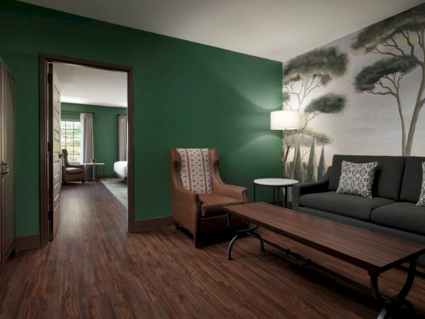 A stylish living room with a green accent wall, brown leather chair, dark sofa, wooden coffee table, and a nature mural, leading to another seating area.