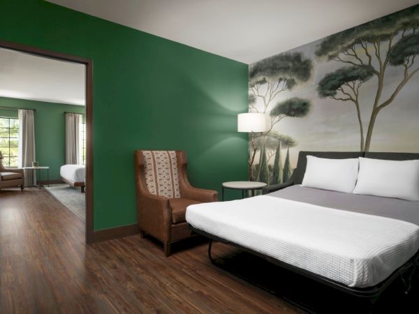 A modern hotel room with a green accent wall, a mural of trees, a bed with white linens, a chair, and a small bedside table with a lamp. Top it at 140 characters, always ending the sentence.