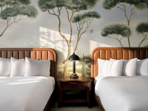 Two twin beds with white linens and wooden headboards sit side by side, a nightstand with a lamp between them, and a mural of trees on the wall.