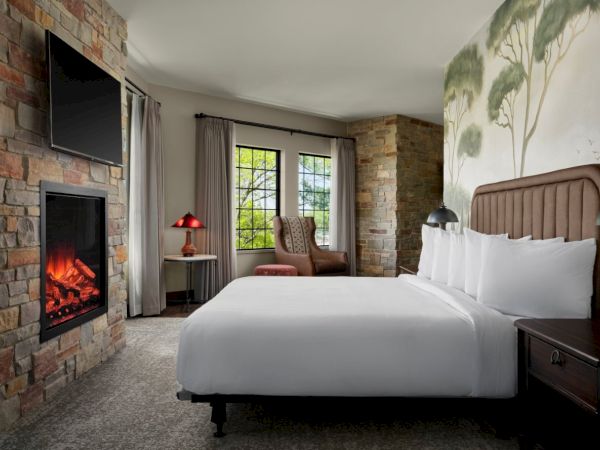 A cozy hotel room with a stone fireplace, flat-screen TV, two bedside tables, a king bed, and a small seating area by a window, bright and inviting.