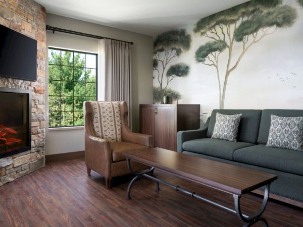 A cozy living room with a stone fireplace, a large window, a patterned mural of trees, a dark green sofa, a brown armchair, and a wooden coffee table.