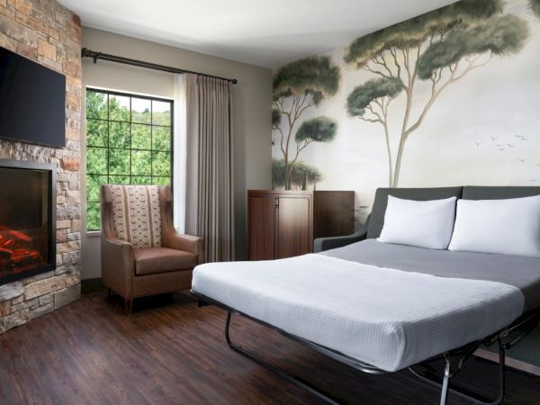 A cozy hotel room with a mural of trees on the wall, a king bed, a stone fireplace, a flat-screen TV, and a brown chair by a window.