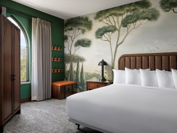 A stylish hotel room with a mural of trees, a large bed with white linens, a wooden headboard, a bench, and a curved-arch doorway by green walls.