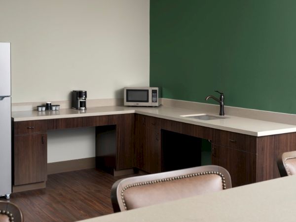 A small office kitchenette with a fridge, coffee maker, microwave, sink, and dark wood cabinets along a green wall, plus a beige countertop and tables.