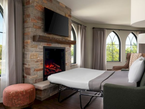 A cozy hotel room with a stone fireplace and mounted TV, a comfy bed, arched windows, and soft curtains for a relaxing retreat.
