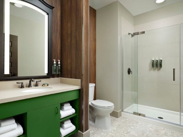 A modern bathroom with a green vanity, white countertop, wall mirror, and glass-enclosed shower. A toilet sits between the sink and shower, towels stocked neatly.