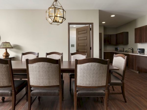 A spacious dining room with a dark wood table and six upholstered chairs, pendant light overhead, open kitchen in the back, warm and inviting.