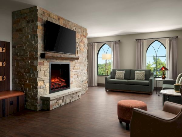 Cozy living room with a stone fireplace and mounted TV, large arched windows, neutral sofa, ottomans, and warm wood floors, sunny and inviting.