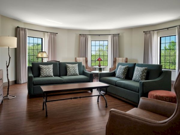 A bright, spacious living room with two dark teal sofas, a matching loveseat, a wooden coffee table, and multiple large windows letting in natural light, cozy throw pillows and a lamp setup, all on warm wooden floors.