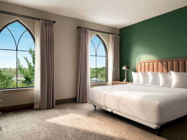 A bright hotel room with two large arched windows, a green accent wall, a white bed with multiple pillows, and neutral curtains. The space looks clean and inviting.
