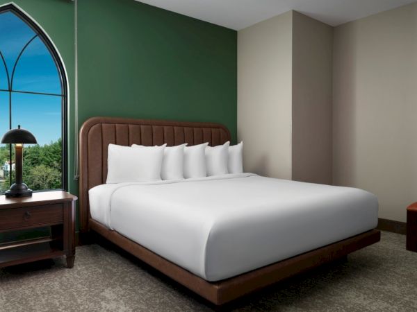 A modern hotel room with a large bed, brown headboard, white bedding, green accent wall, and an arched window beside a nightstand with a lamp.