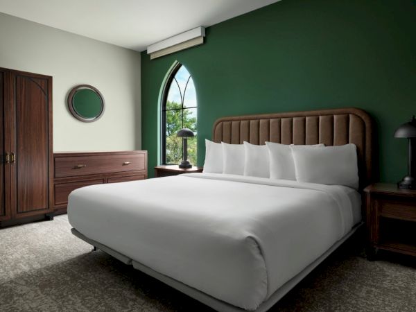 A modern hotel bedroom with a king bed, dark wood furniture, green accent wall, arched window, and neutral carpet.