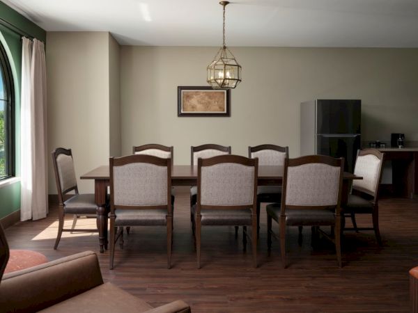 A formal dining room with a long wooden table and eight upholstered chairs, a hanging light fixture, large window with curtains, and a sideboard against a beige wall.