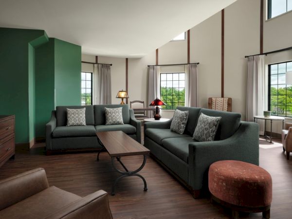 A spacious living room with green accent wall, two grey sofas, a wooden coffee table, armchairs, red ottoman, and large windows letting in natural light.