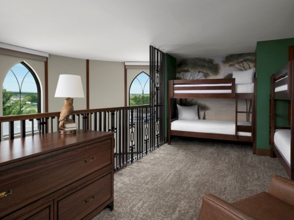 A spacious dorm-style hotel room with bunk beds, a dresser, arched windows, and a seating area; warm wood furnishings and a balcony railing.