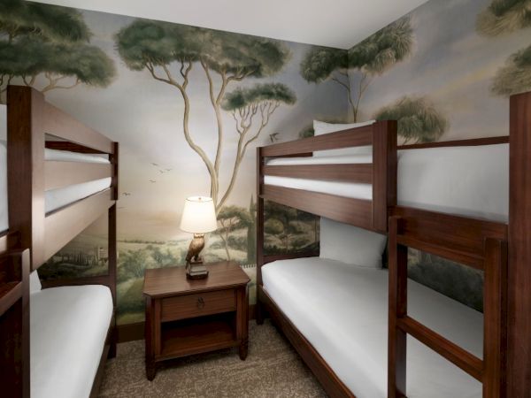 A small dorm-style room with four wooden bunk beds, a nightstand with a lamp, and a nature mural of trees on the walls.