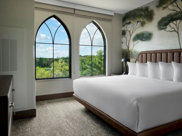 A modern hotel room with a large bed, arched windows overlooking greenery, a mural wall with trees, and neutral decor, bright and serene.