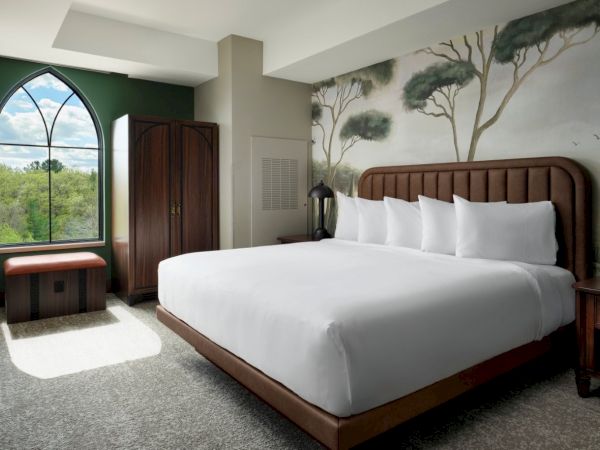 A stylish hotel room with a large bed, mural headboard, wooden furniture, and a green window offering a serene view of trees outside.
