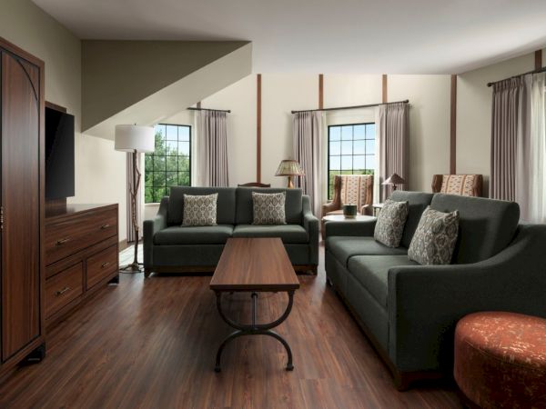 A cozy living room with two dark-green sofas, a wooden coffee table, windows with curtains, and warm wood flooring.