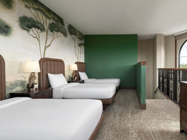 A hotel room with two single beds, nightstands, lamps, a nature mural, a green accent wall, and a balcony railing by large windows.