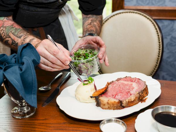 A chef with tattooed arms plates carved steak and mashed potatoes, garnishing with microgreens using tongs at a table.