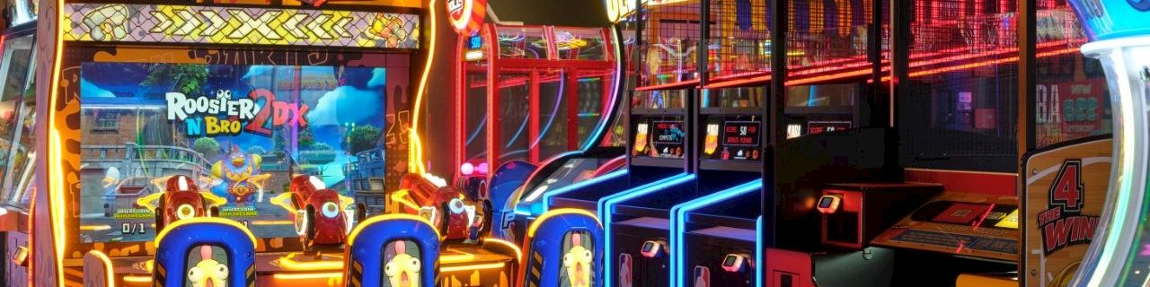 A brightly lit arcade with &ldquo;Rooster N Bro 2DX&rdquo; shooting games and colorful prize-style machines.