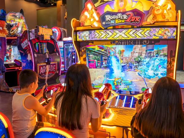 Two kids play a neon-lit arcade shooter game, firing at targets on the screen inside a prize arcade.