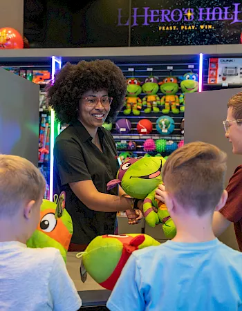 A smiling toy-store worker hands a green plush dinosaur to children at an arcade, with colorful games and signs behind her.