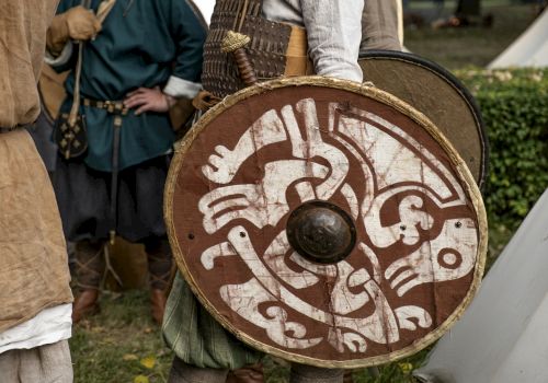 A medieval reenactor holds a round shield with white painted designs while wearing chainmail and a conical helmet.