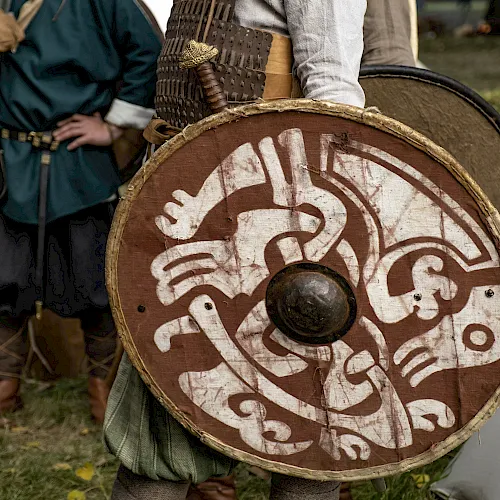 A medieval reenactor holds a round shield with white painted designs while wearing chainmail and a conical helmet.