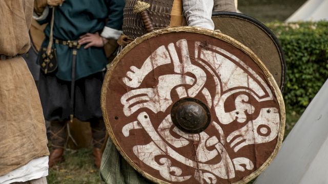 A medieval reenactor holds a round shield with white painted designs while wearing chainmail and a conical helmet.