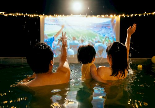 Two swimmers in a pool cheer as rain and confetti fall, watching a big concert screen under stadium lights.
