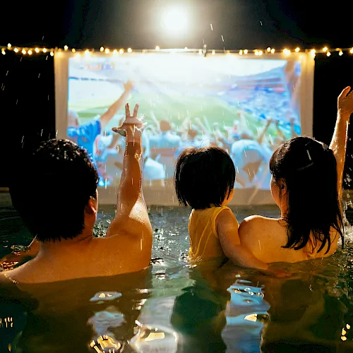 Two swimmers in a pool cheer as rain and confetti fall, watching a big concert screen under stadium lights.