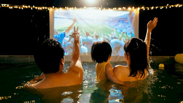 Two swimmers in a pool cheer as rain and confetti fall, watching a big concert screen under stadium lights.