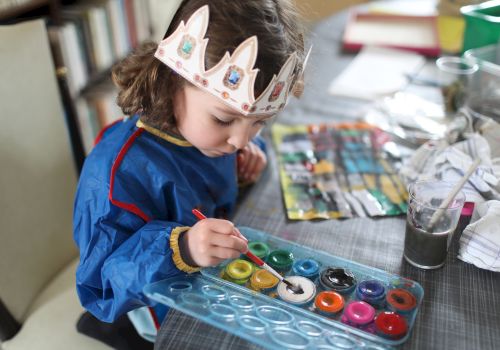 A child in a colorful crown headband paints at a table with brushes, paint pots, and a cup of water.