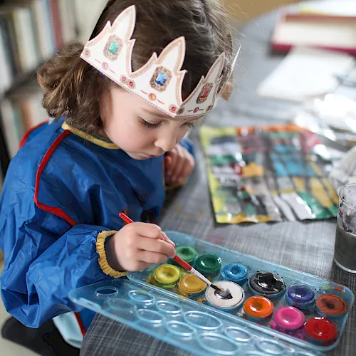 A child in a colorful crown headband paints at a table with brushes, paint pots, and a cup of water.