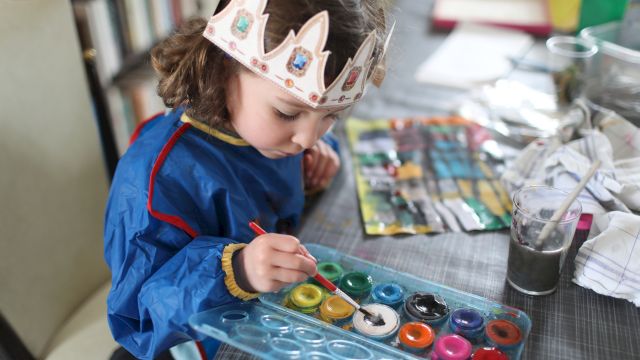A child in a colorful crown headband paints at a table with brushes, paint pots, and a cup of water.