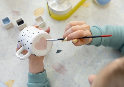 A child paints a white ceramic mug with a brush, adding face details while crafting at a table.