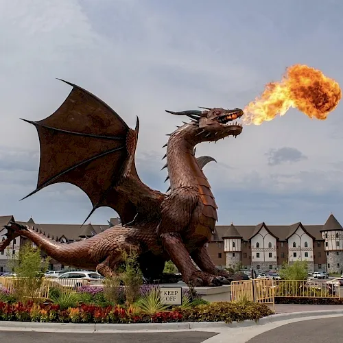 A giant cartoon-style dragon breathes fire over a landscaped hotel entrance with cars and buildings in the background.